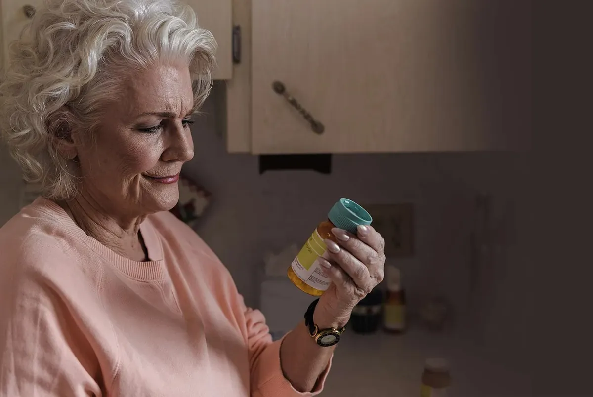 A woman reading the label of a supplement bottle in a kitchen